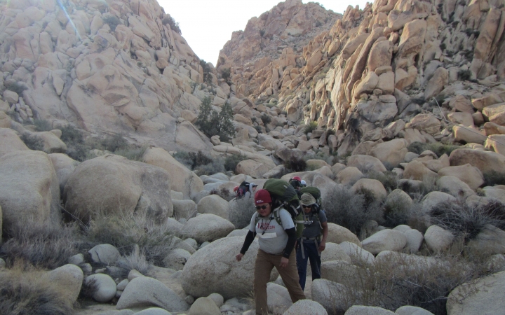 A group of people wearing backpacks make their way through an area populated by large rocks.